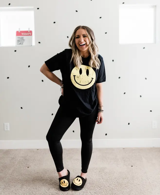 Dani, founder of the Bestie Squad, standing in a black smiley-face tee and smiley slippers in front of a polka-dot wall.