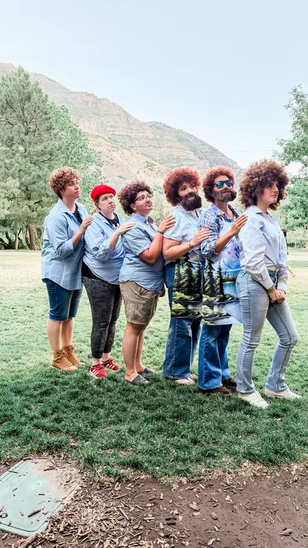 Bestie Squad members in curly wigs and costumes striking a playful pose on the grass.
